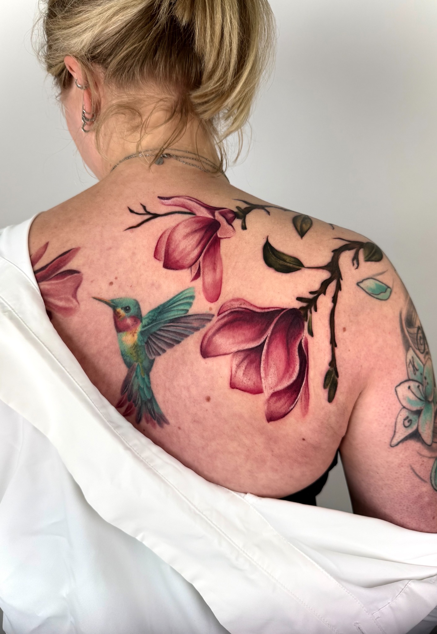 Farbiges Tattoo mit einem Kolibri und großen rosafarbenen Magnolienblüten auf dem oberen Rücken