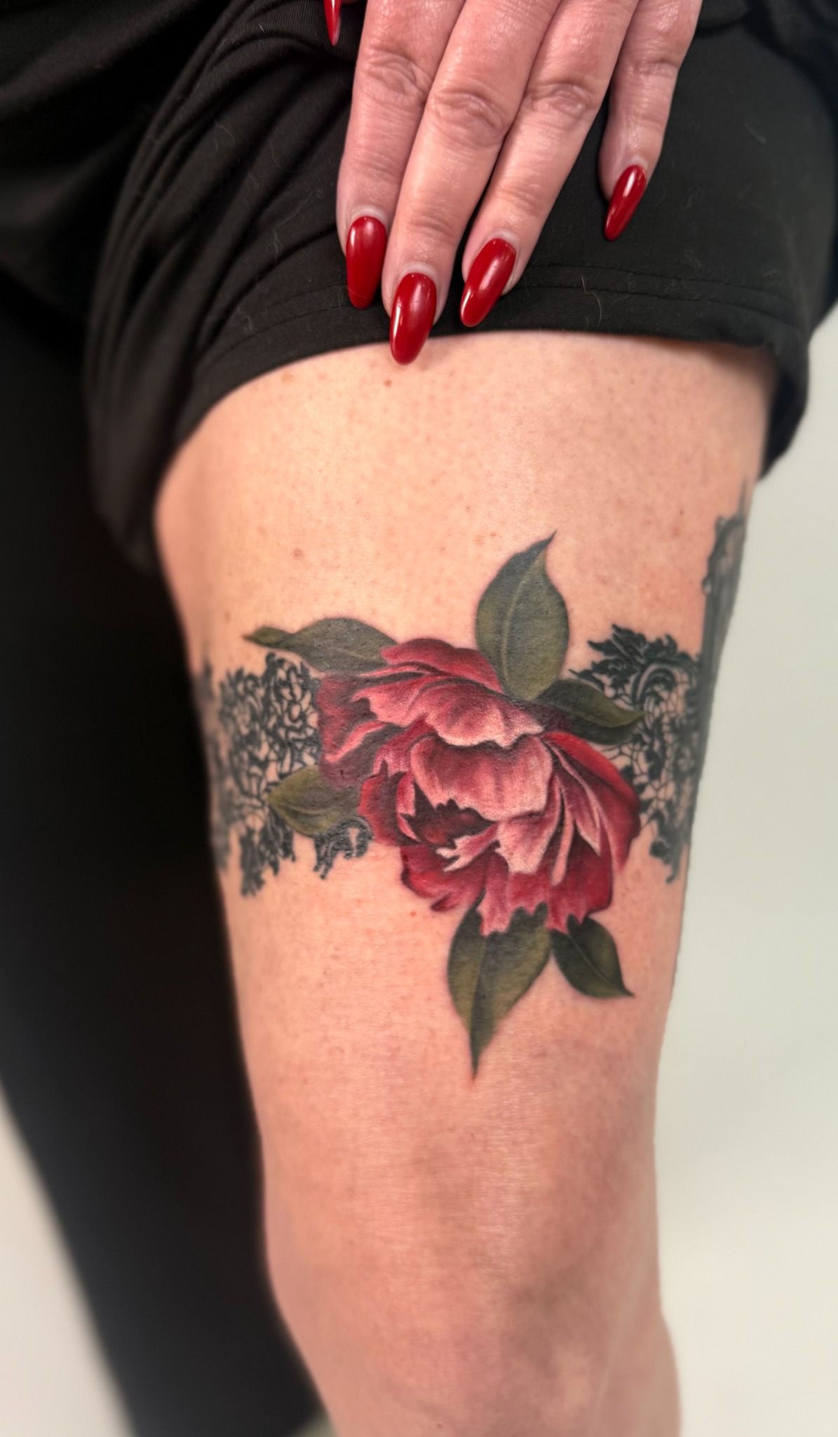 Rote Pfingstrosenblüte mit grünen Blättern und dunklen Details als Tattoo am Oberschenkel