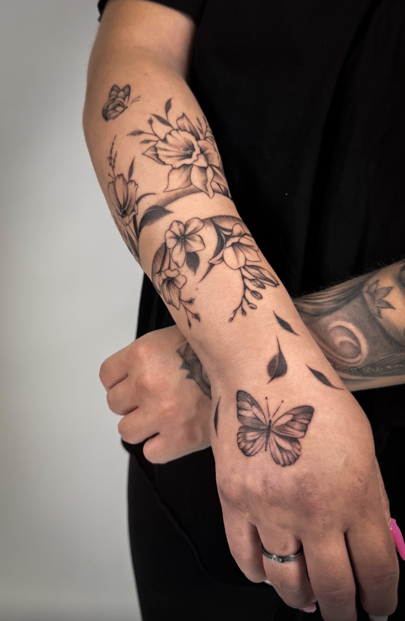 Fine Line Tattoo mit Blumen am Unterarm und einem Schmetterling auf dem Handrücken