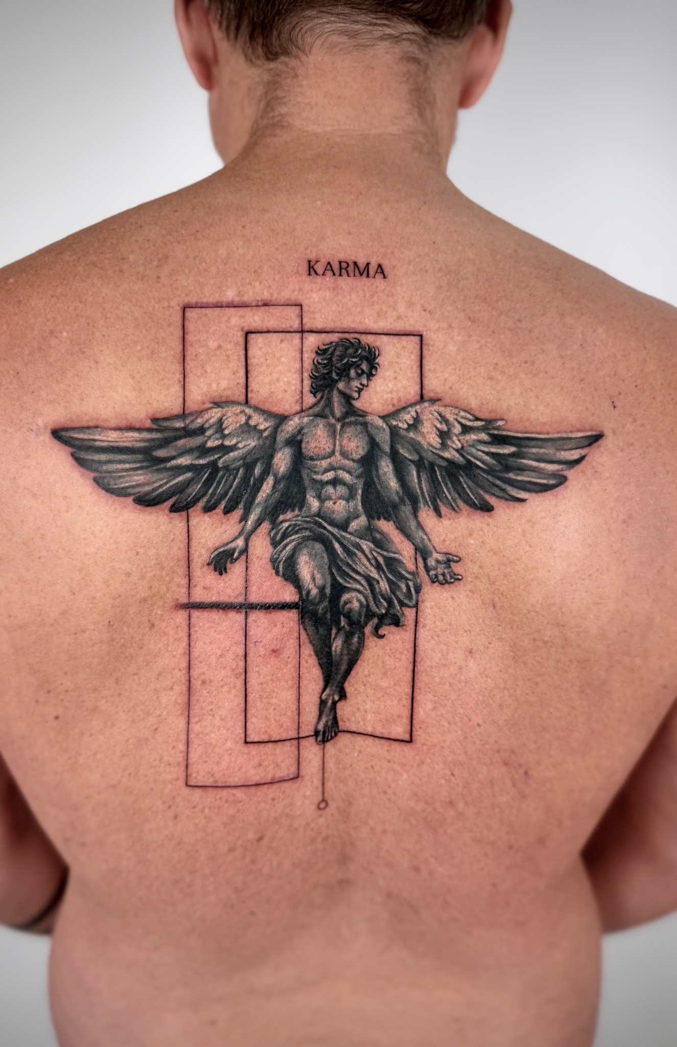 Black & Grey Tattoo einer geflügelten Figur auf dem Rücken mit geometrischen Rahmen und dem Schriftzug Karma