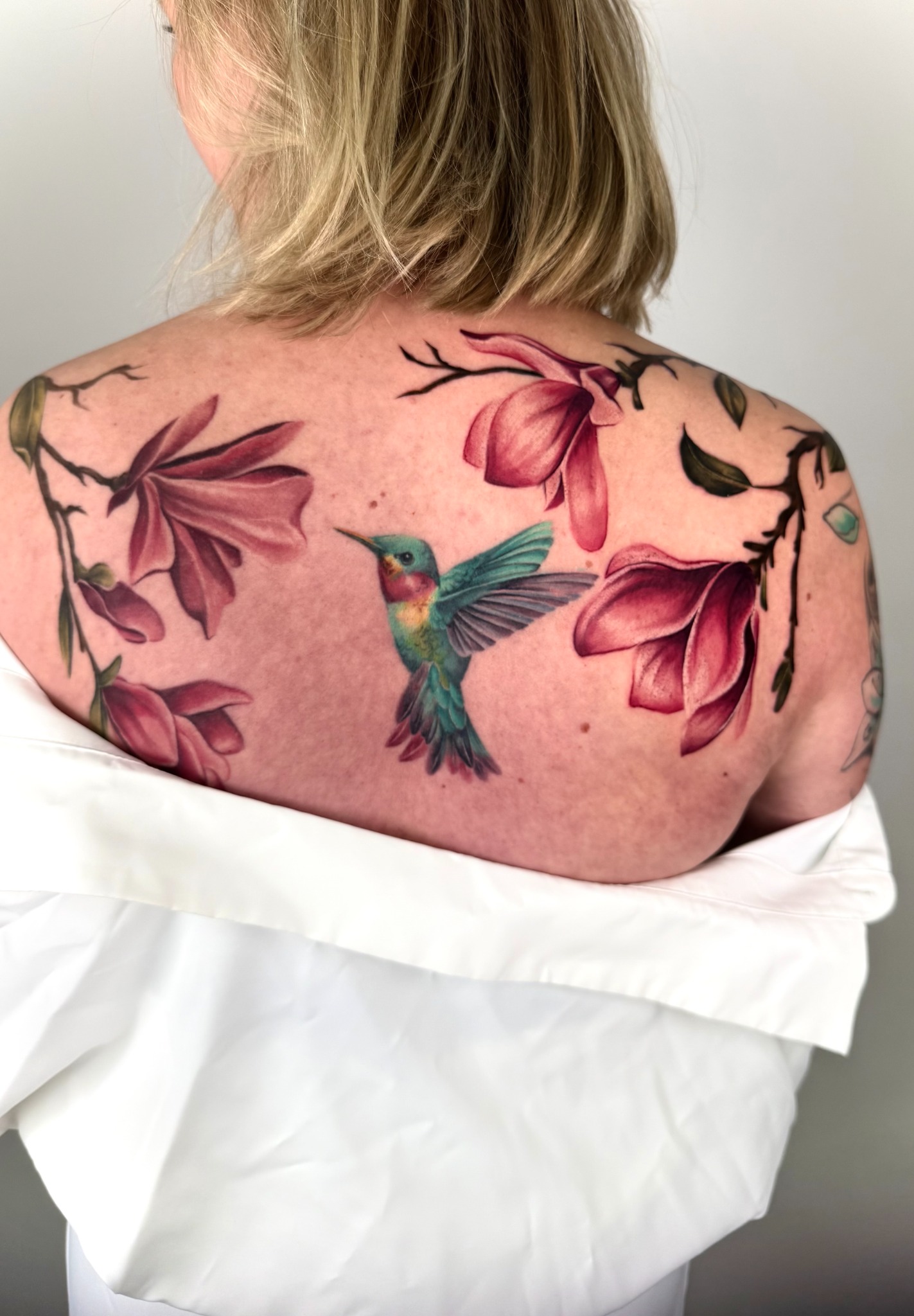 Farbiges Tattoo mit grünem Kolibri und rosa Magnolien auf dem oberen Rücken
